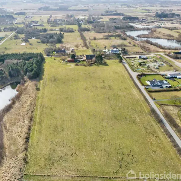 En stor, grøn mark strækker sig i et åbent landskab med få træer. I baggrunden ses spredte bygninger og veje. En sø er placeret til venstre.