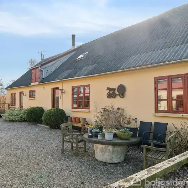 Et gult hus med røde vinduer står i en grusbelagt gårdsplads. Foran huset er der bænke og et bord med blomsterkrukker. Baggrunden viser træer og en klar himmel.
