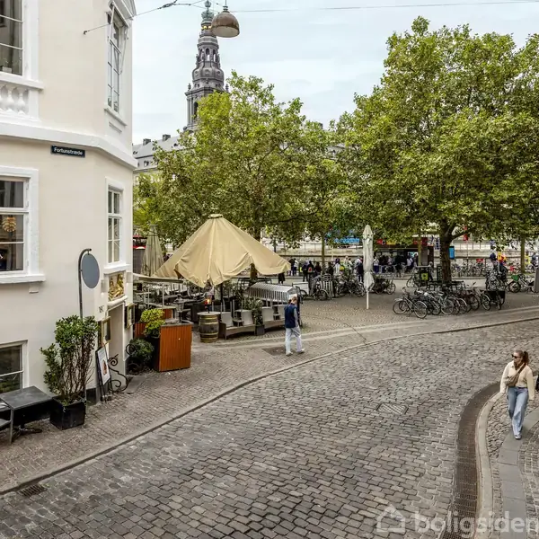 Gaden er belagt med brosten og omgivet af bygninger med butikker. To mennesker går langs fortovet. En café med terrasse ligger på hjørnet, og der er træer i baggrunden.