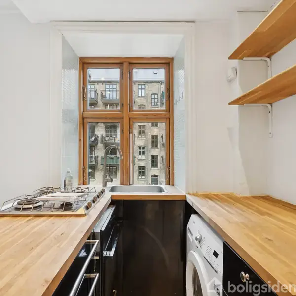 Køkken med træbordplader, gaskomfur og vaskemaskine integreret. Store vinduer viser en beboelsesejendom med dekorative facader. Til venstre ses en glasdør, og øverst er der hylder.