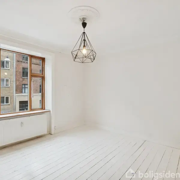 Et tomt rum med en moderne, geometrisk lysarmatur hængende fra loftet. Væggene og gulvet er hvide. Et stort vindue lader naturligt lys strømme ind og afslører en udsigt til nabobygningen.