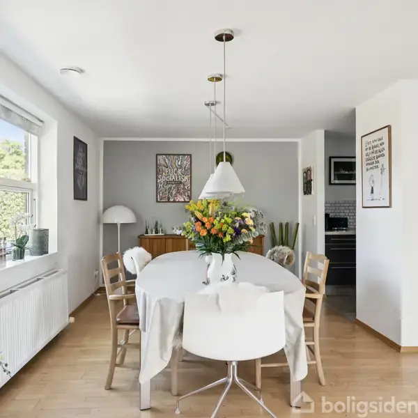 Et ovalt spisebord med en vase blomster står i et lyst spisestueområde. Omkranset af stole og kunst på væggene. Store vinduer giver naturligt lys.