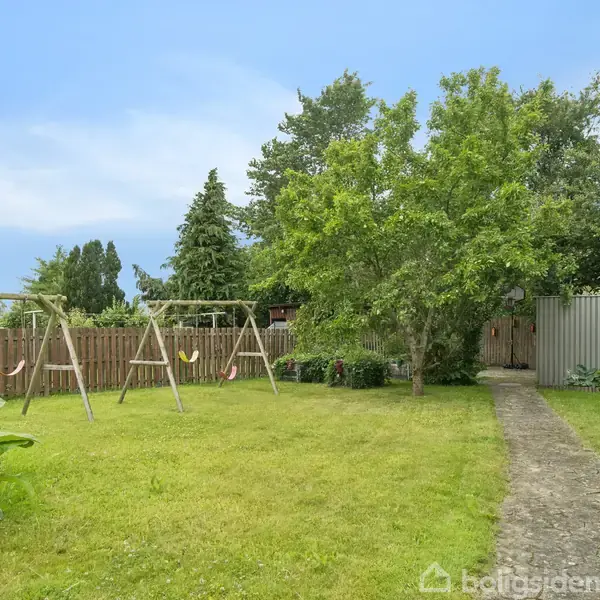 Gyngestativ står stille på en græsplæne omgivet af buske og træer. En smal sti fører gennem haven mod et trægærde under en blå himmel.