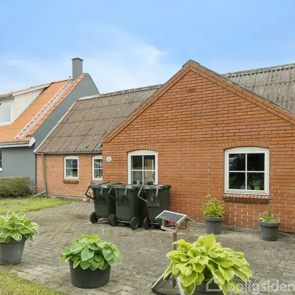 Et rødt murstenshus med skråt tag har skraldespande placeret foran. I haven er der flere store potteplanter. Nabohuset har et gråt tag og en hæk adskiller de to huse.