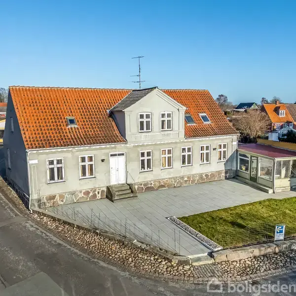 Et gråt hus med rødt tag står ved en vej. Det har en veranda og små vinduer på første sal. Huset er omgivet af flisebelægning og en lille græsplæne.