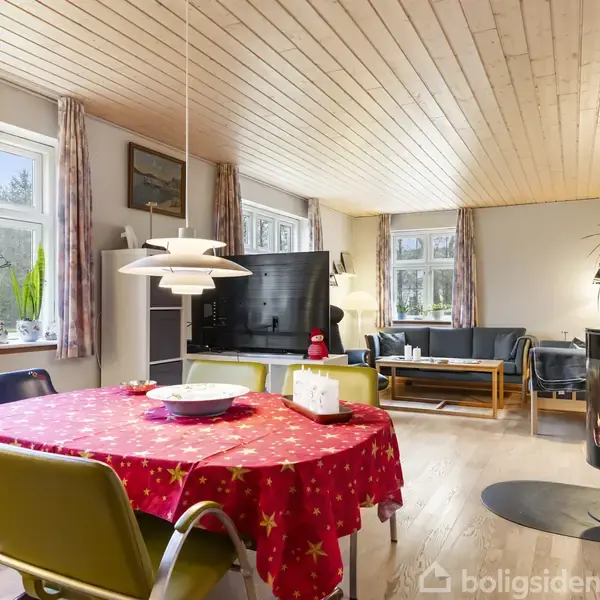 Spisebordet er dækket med en rød dug og omgivet af stole, placeret i en lys stue med trægulv og træloft. Der er også en brændeovn og sofaarrangement.