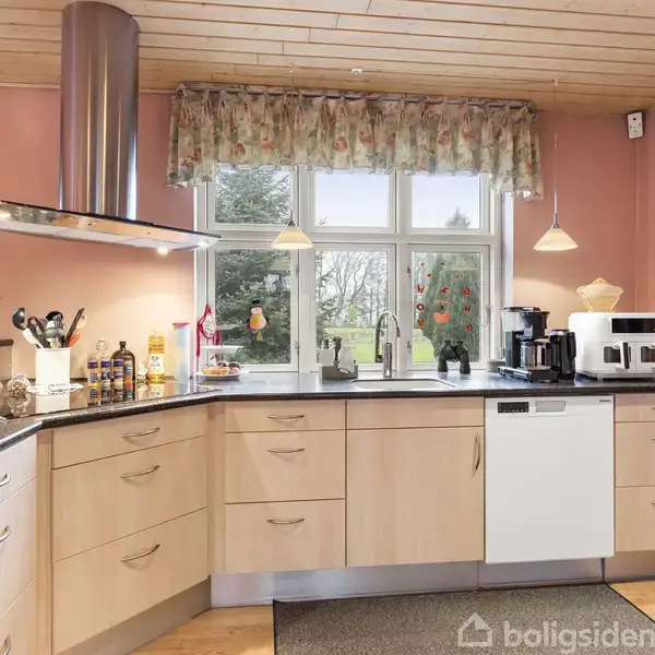 Et rummeligt køkken med lyse træskabe og sort bordplade står i en hyggelig atmosfære. Der er en kaffemaskine, opvaskemaskine og diverse køkkenartikler. Udsigt til en have gennem et stort vindue.