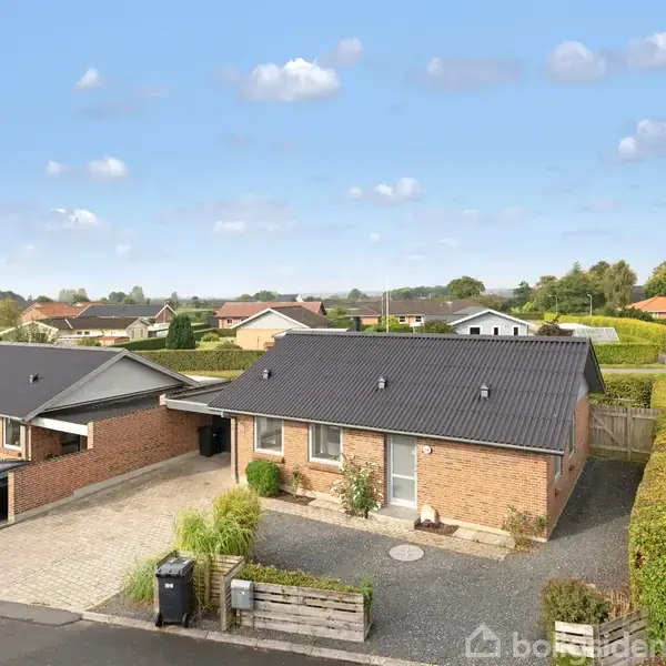 To ens, murstenshuse med sorte tage står i et parcelhuskvarter. Bilen holder i indkørslen, omgivet af hæk og flisebelagt vej. Skyfri blå himmel i baggrunden.