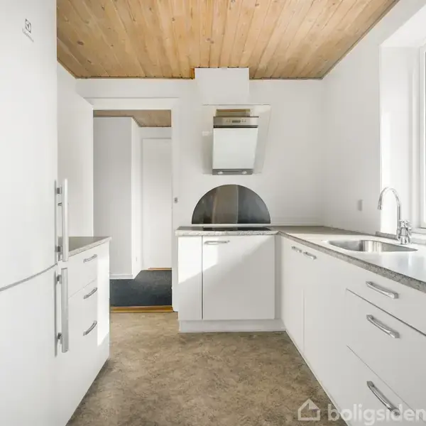 Et moderne, hvidt køkken med stålvask og komfur. Køleskabet står til venstre, og der er en emhætte over kogepladerne. Rummet har lyst træloft og vindue mod højre.