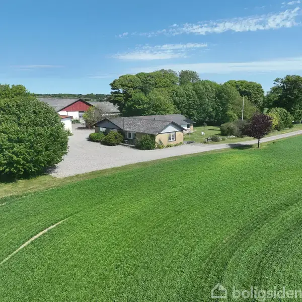 Et hus står omgivet af en stor, grøn mark og træer. Ved siden af huset ses en rød lade og en grusvej. Himlen er blå med få skyer.