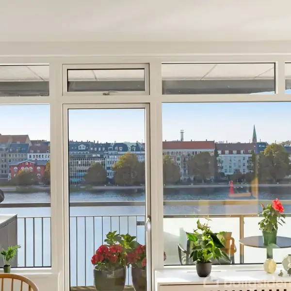 En stue med store vinduer viser en udsigt over en flod og bygningsrække i baggrunden. Indenfor er der planter i potter og nogle møbler ved siden af et spisebord.