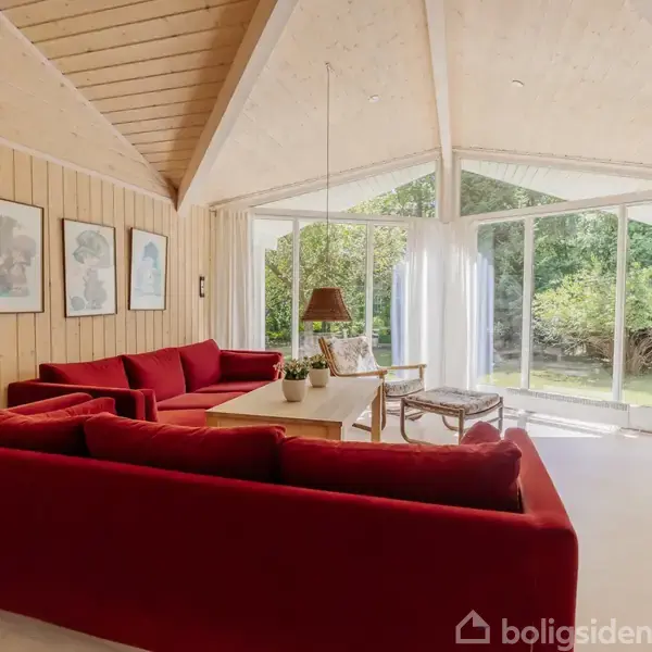 Rød sofa i stue placeret ved stort vindue, der giver udsigt til en have. Væggene har lyse træpaneler og dekoreret med tre malerier.