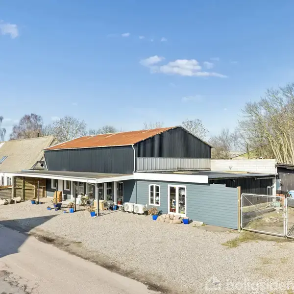 Et stort, moderne hus med en grå facade og rødt tag. Bygningen står på en grusbelagt grund omgivet af træer under en klar blå himmel.