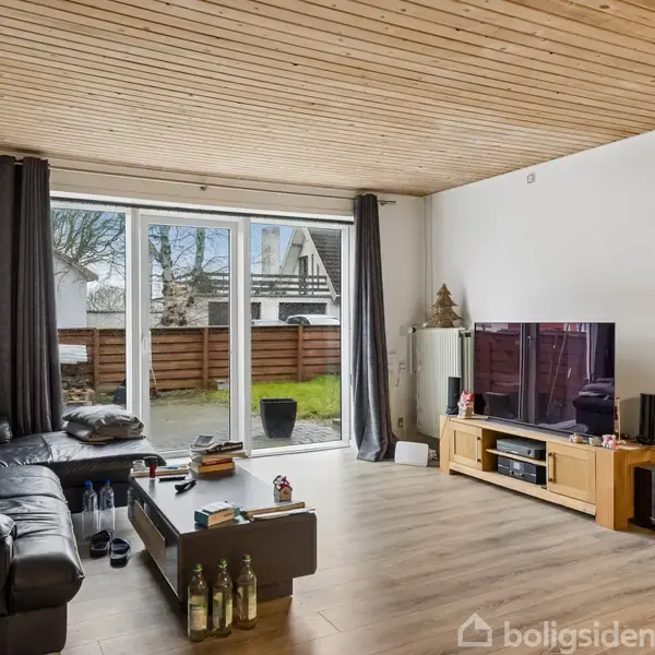 Sort lædersofa i hjørnet; rummet med trægulv og -loft. Foran sofaen, et sofabord med flasker og glas. Stor fladskærms-tv på tv-bord. Store vinduer fører til terrasse med græsplæne.