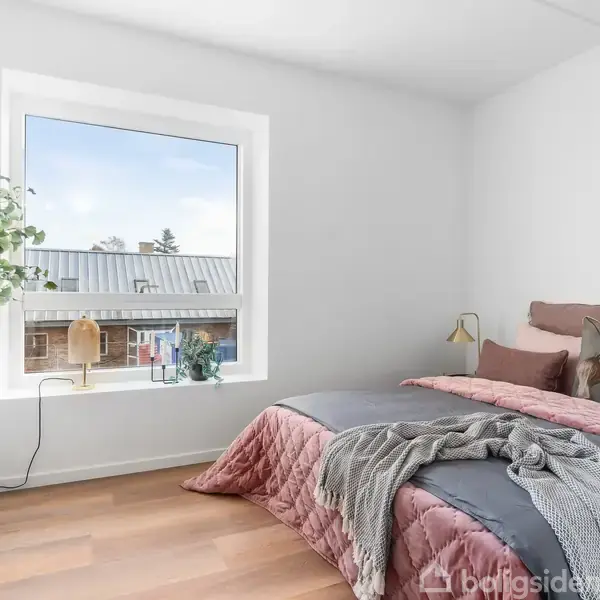 Soveværelse med en seng dækket af grå og rosa sengetøj. En lampe er fastgjort til væggen. Et vindue viser udsigt til hustage. En plante står i hjørnet.