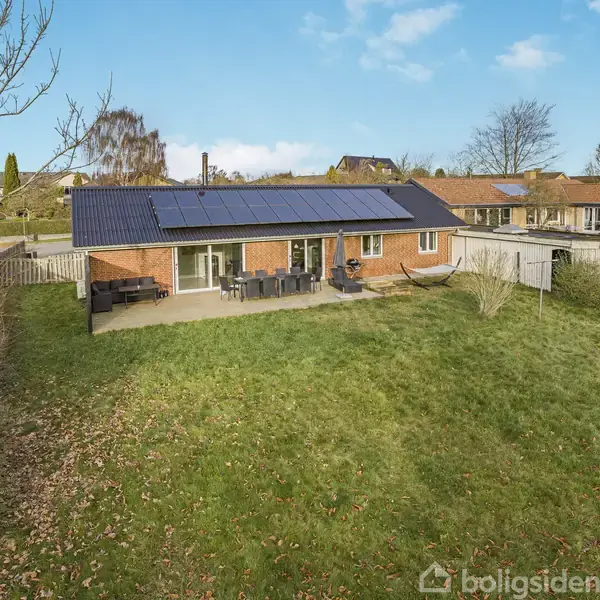 En murstensvilla med solpaneler på taget står i en grøn have med terrasse. Haven er omgivet af træer, og der er en havelåge i træhegn. Himlen er blå.