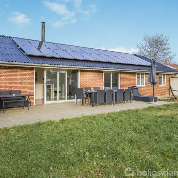 Rødstenshus med solpaneler på taget, stående i en have med græsplæne. En terrasse med sofa, bord og stole er synlig, under en blå himmel med få skyer.