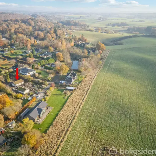 Rødt hus markeret med en pil står blandt andre huse. Omkringliggende landskab består af marker og skov. Byen ligger i et åbent, landligt område.