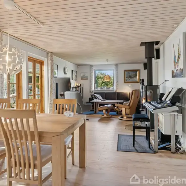 Træspisebord med stole står i en lys stue. Der er glasdøre til haven, sofa, pejs og et klaver i rummet. Væggen har malerier, og loftet er i træ.