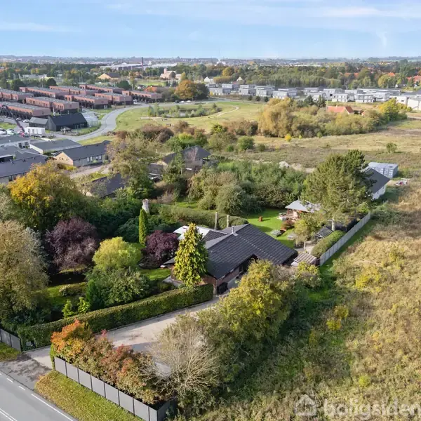 Et hus omgivet af træer og buske ses fra luften. Boligen ligger i et beboelsesområde med flere huse tæt på, og marker strækker sig i baggrunden.