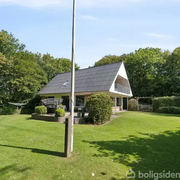 Et hus står stille i en velplejet have, omgivet af træer. Der er en flagstang foran huset og et legeområde med en rutsjebane til venstre.