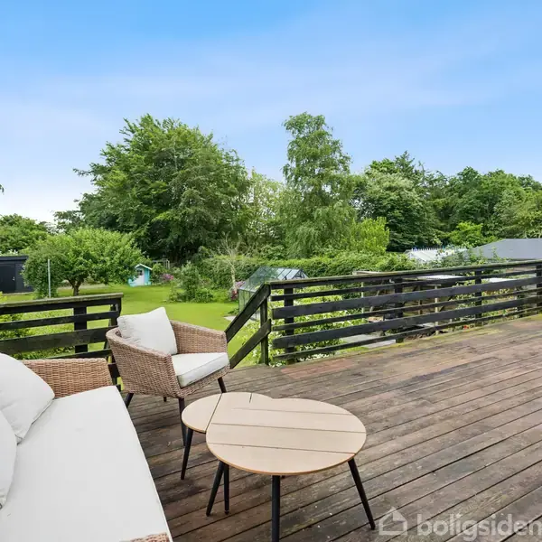 En terrasse med trægulv har en hvid sofa og et lille, rundt bord. I baggrunden er der en frodig have med græsplæne og træer under en klar blå himmel.