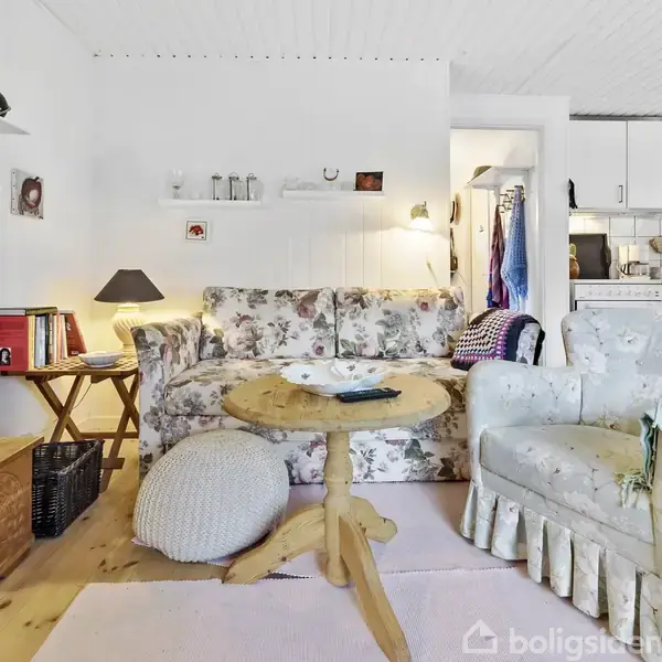 En stue med en blomstrede sofa og lænestol omkring et rundt træbord. En lampe og tv står på sidebordet. Baggrunden viser et køkken med hvide skabe.