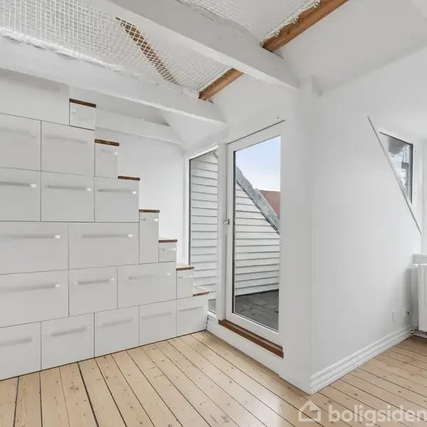 Trappe med skabsopbevaring fører opad, placeret i lys rum med trægulv. Store vinduer og hvidmalede vægge skaber en luftig atmosfære. Dør til altan ses på højre side.