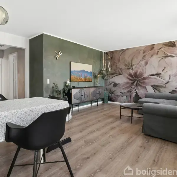Stue med spisebord dækket med dug og sorte stole i forgrunden, grå sofa ved siden af blomstret vægdekoration i baggrunden, moderne indretning og træpanelgulve.