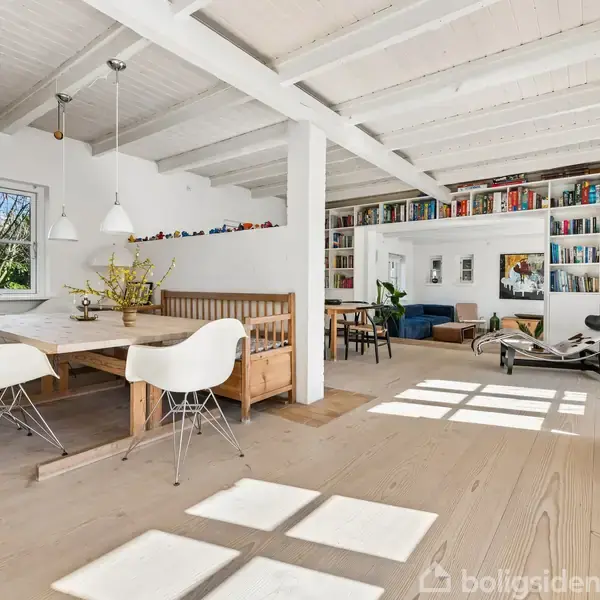 Lyst rum med træbord og hvide stole omkring det. Baggrunden viser en sofa, reoler med bøger, og stort vindue. Hvide bjælker pryder loftet, og der er dekorative planter.