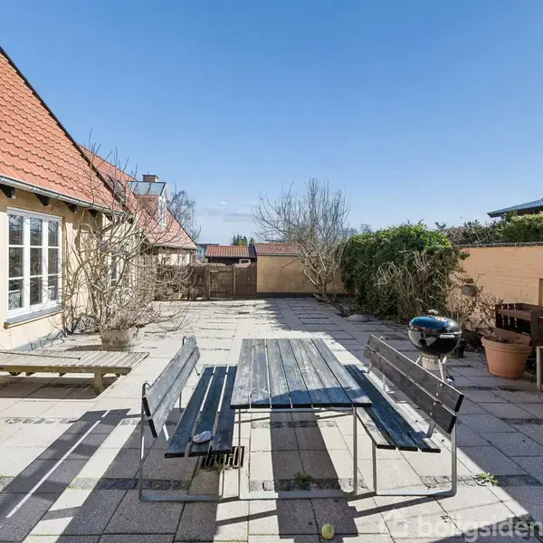 En lang træbænk med et bord står på en flisebelagt terrasse. Omkring er der gule vægge, rødtagede bygninger, og grønne buske. En grill står til højre side.