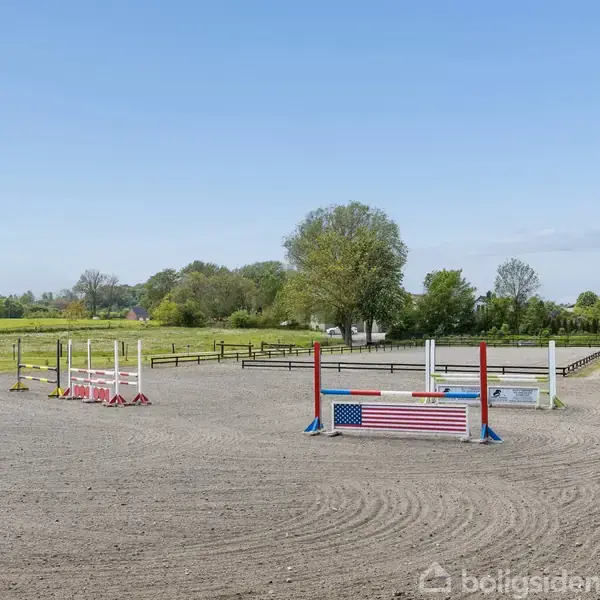 Ridebane med springforhindringer. Bander i baggrunden afgrænser det træomkransede felt. Grønt græs og træer omkranser området, der ligger under en blå himmel.