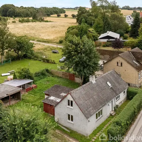 En landsbyhus omgivet af grønne træer og marker står langs en vej. I baggrunden ses en stor græsplæne med et lille skur. Unavngivet vej fører gennem landskabet.