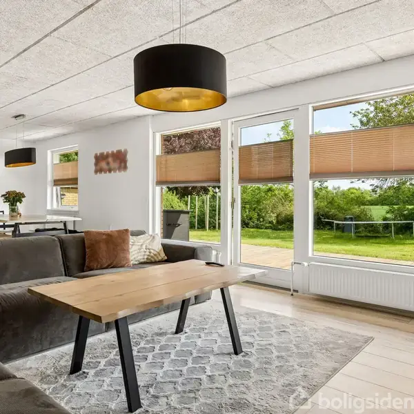 Stor, komfortabel sofa står foran sofabord i stue med store vinduer og udsigt til grøn have. Rundt spisebord i baggrunden. Lysekroner hænger fra loftet.
