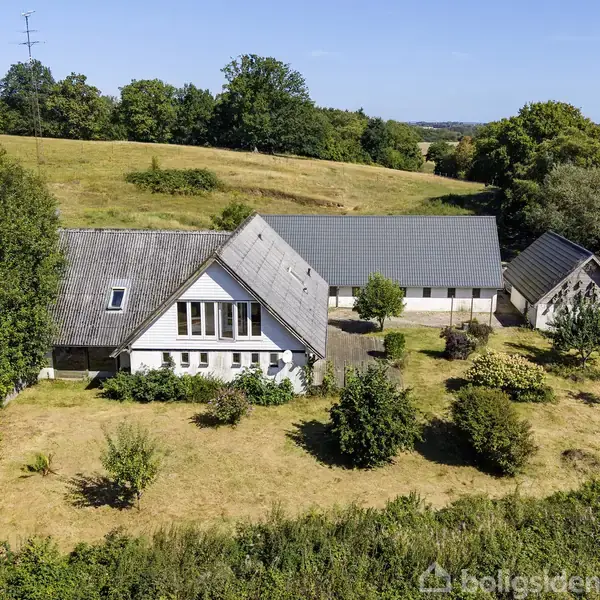 Et hvidt hus med grått tag står i en åben, grøn landskab med træer omkring. Det ser landligt ud med omkringliggende marker og bakker i det fjerne.
