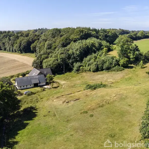 En gård med gråtagede bygninger ligger stille omgivet af åbne marker og tæt skov i et landligt område under en klar blå himmel.