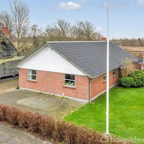 En rødstensbungalow står på en grøn græsplæne, omkranset af buske. Den har mørkt tag og flere vinduer. Foran huset er en flisebelagt indkørsel. Omkringliggende træer ses i baggrunden.