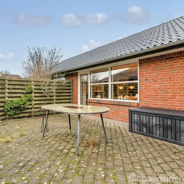 Et rundt bord står på en flisebelagt terrasse ved siden af en rød murstensbygning. En sort bænk er placeret ved væggen. Træhegn og buske omkranser området.