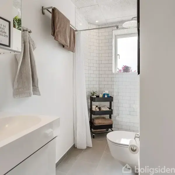Badeværelse med hvidt håndvaskskab, bruseområde med forhæng og stort vindue. Hylde med planter og håndklæder. Skilt på væg: "Relax, it's just a phase."