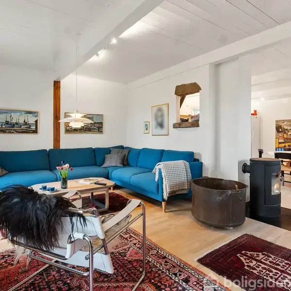 Blå sofa står i en stue med malerier på væggen. En brændeovn står tæt på et bord med stole. Tæpper dækker dele af trægulvet.