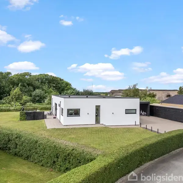 Hvidt, moderne hus står i en velplejet have med hække og græsplæne. Omkringliggende område består af flere huse og træer under en blå himmel med få skyer.