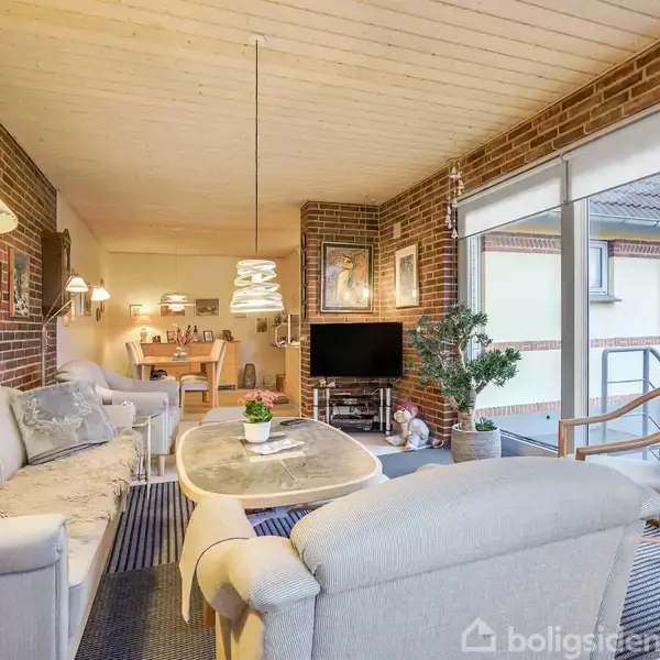 Stue med lyse møbler omkring et sofabord, TV på et skab ved siden af store vinduer, der afslører en terrasse. Omkring er der murstensvægge og dekorative lamper.