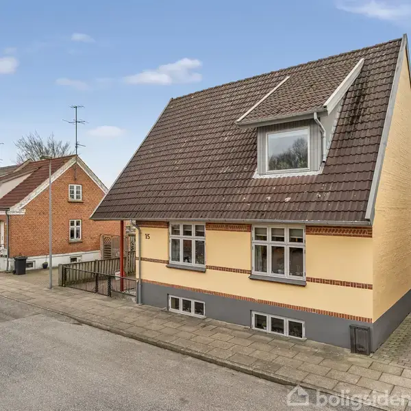 Et gult hus med brun tag og dannebrogsvinduer står ved en stille gade med fortov. Til højre ses en carport med en parkeret blå bil. Nummeret 15 ses på husvæggen.
