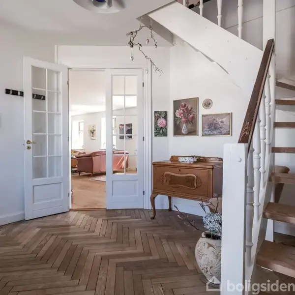 Trappe leder opad i en entré. Der er et træskab mod væggen med blomsterdekorationer. En åben dør i baggrunden fører til en stue med sofaer.