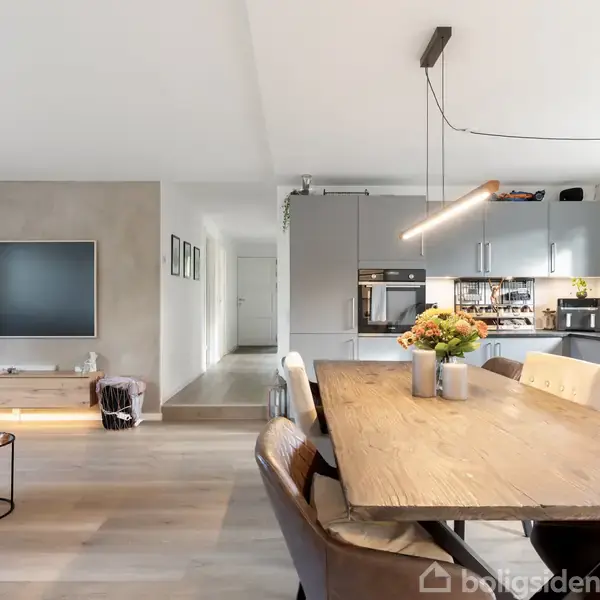 Spisebord med blomster på står centralt, omgivet af stole i en moderne stue-køkken kombination. Fladskærms-TV på væggen, køkken i baggrunden med grå skabe og naturligt lysindfald.