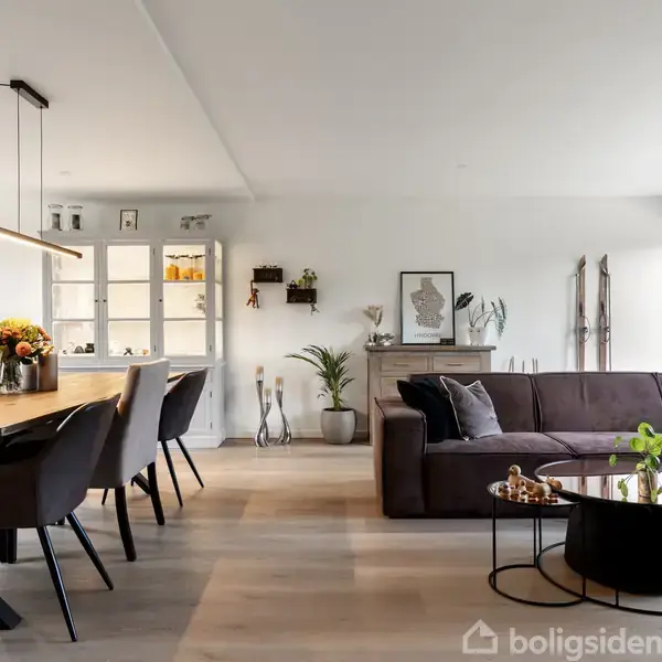 En stue med en stor brun sofa står centralt; sofabord med planter og dekorationer foran. Ved siden af står et spisebord med stole. Væggene har hylder og dekorationer.