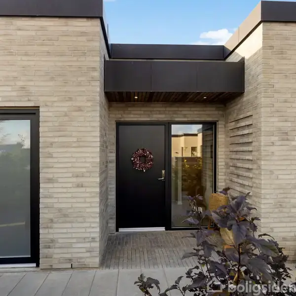 En moderne bygning har en sort hoveddør dekoreret med en krans. Omkring døren er murstensvægge. I forgrunden er en plante og bagved er en glaskuppel på væggen.