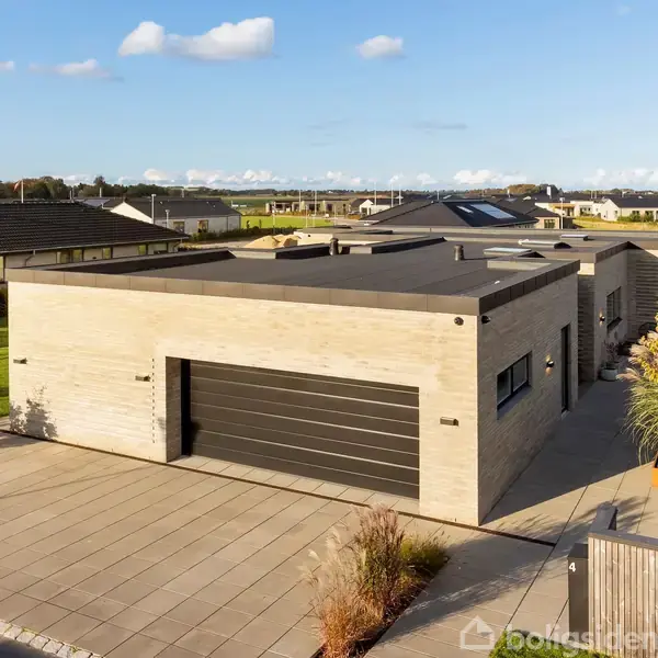 En moderne fladtagsejendom står på en brolagt indkørsel, omgivet af velplejet græsplæne og blomsterbede. I baggrunden ses et boligområde under en klar blå himmel med spredte skyer.