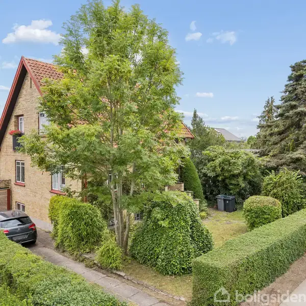 Et murstenshus med rødt tag står i en velplejet have omgivet af hække og træer. En bil er parkeret ved indkørslen, og huset ligger i et villakvarter.