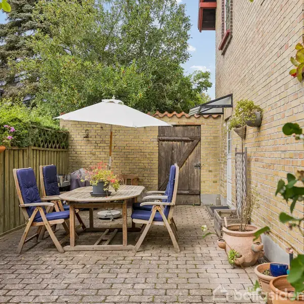 En rund træbord med blå polstrede stole står under en parasol på en brostensbelagt terrasse. Omkranset af murstensvæg og træhegn med blomsterkrukker og grønne planter.